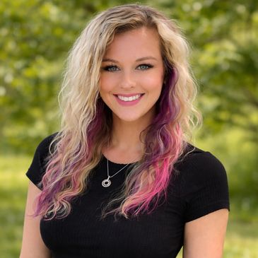 Smiling woman with blonde and pink wavy hair outdoors.
