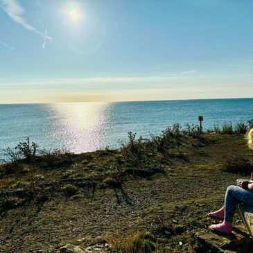 Person sitting near a cliff overlooking the sunlit ocean under a clear sky.