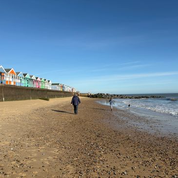 A person walks alone on a sandy beach near colorful beach huts under a clear blue sky.