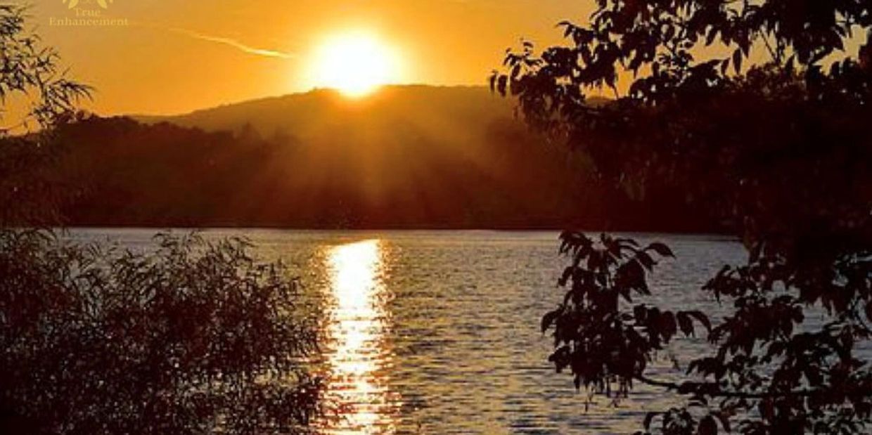 Sunset over a lake with silhouetted trees and hills in the background.