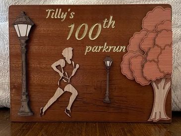 Wooden plaque celebrating Tilly's 100th parkrun with a runner, lamp posts, and tree.