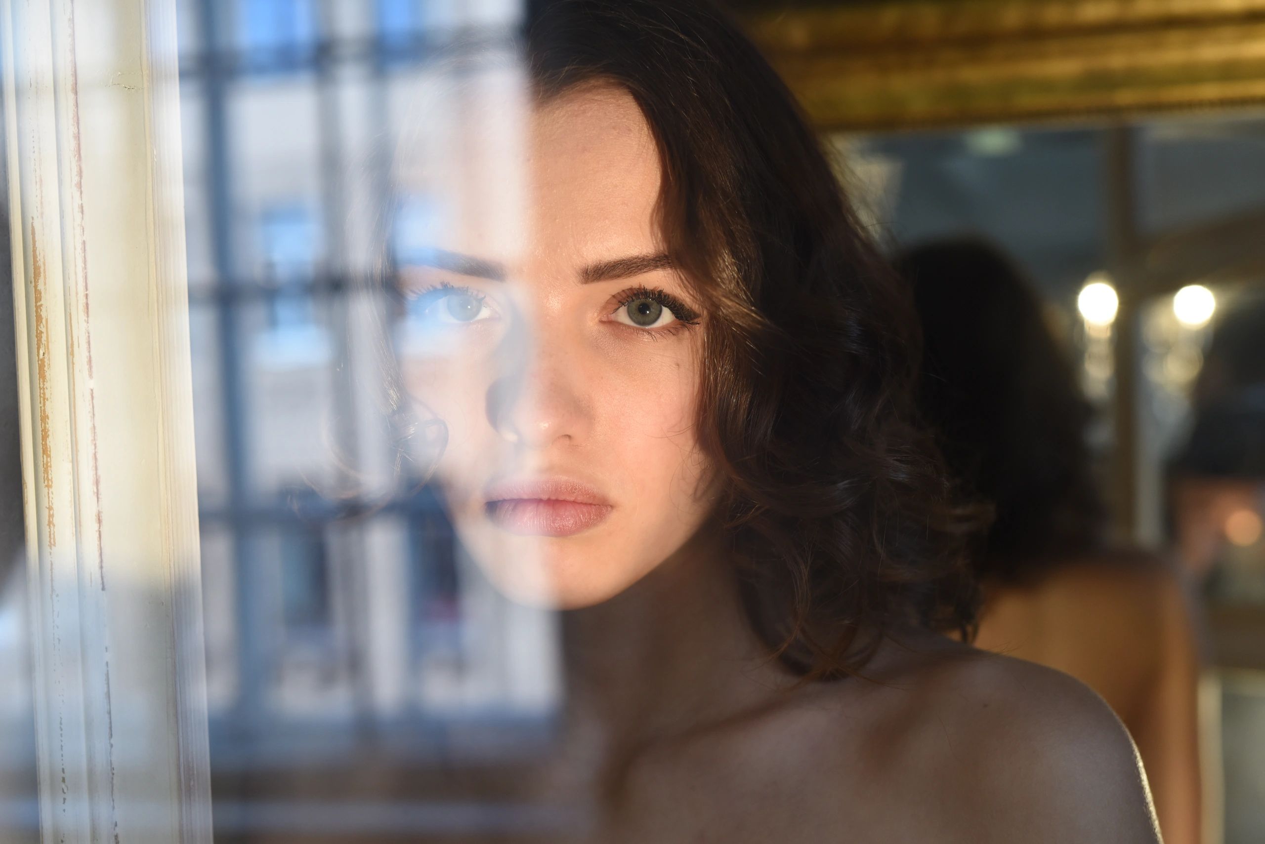 A woman with curly hair gazes thoughtfully through a window with reflections.