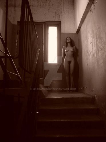 Nude woman stands in dimly lit, worn stairwell near a window.