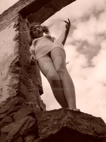 Low-angle sepia photo of a woman standing on a stone ledge near a rough wall.