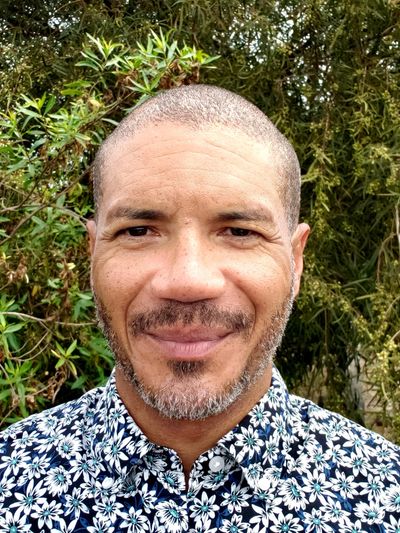 Smiling man in a floral shirt stands outdoors with greenery in the background.