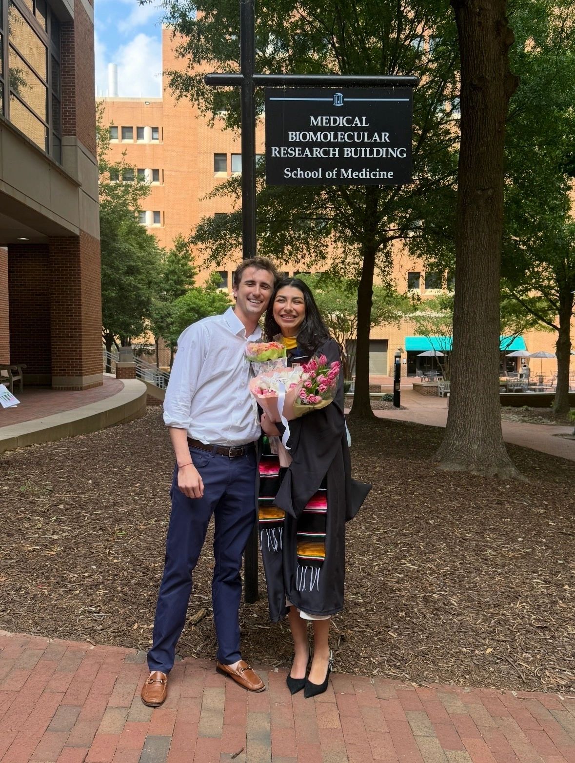 Two people smiling in front of a Medical Biomolecular Research Building sign, one in graduation attire holding flowers.