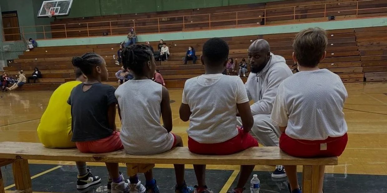 A coach talks to young basketball players sitting on a bench in a gym.