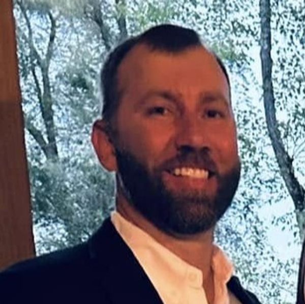 Smiling man in a suit with a forest background.