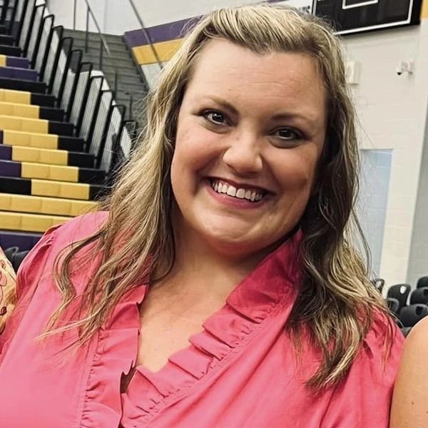 Smiling woman in a pink blouse at a gymnasium with tiered seating.