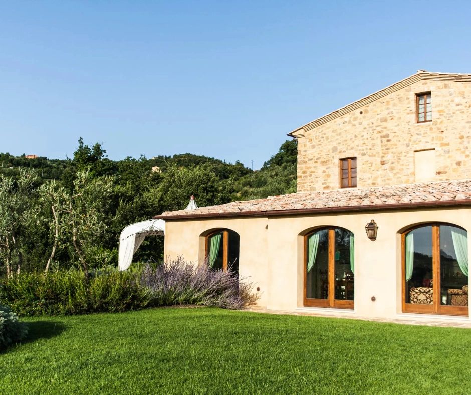 A rustic stone house with large arched windows and a sloped roof, set in a lush green landscape with olive trees and lavender plants.