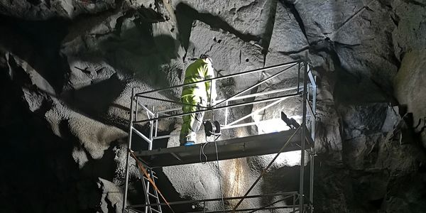 Worker in protective gear on scaffolding inside a rocky cave.