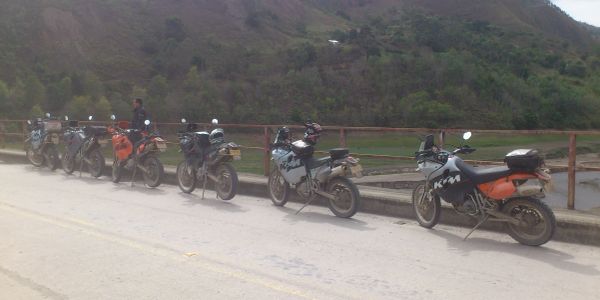 Seven motorcycles parked by a roadside with hills in the background.