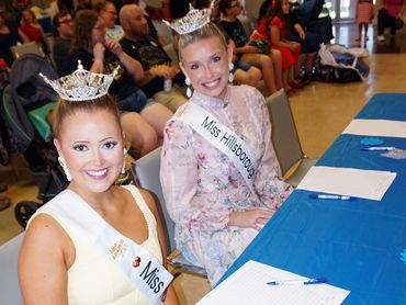 Two smiling beauty queens wearing crowns and sashes at a judging table.