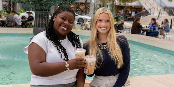Two women smiling and holding iced coffee drinks near a fountain.