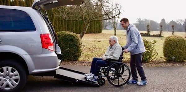 A man helps an elderly person in a wheelchair onto a van ramp.