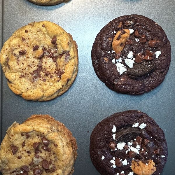Four freshly baked cookies on a baking tray, two classic chocolate chip and two chocolate cookies with Oreo and chocolate chips.