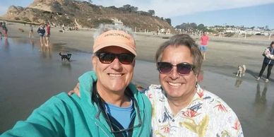 Two men smiling for a selfie on a sunny beach with people and dogs in the background.