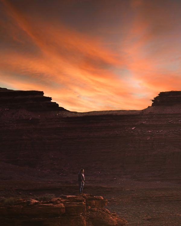 Person standing on rocky cliff at sunset with vibrant orange sky.