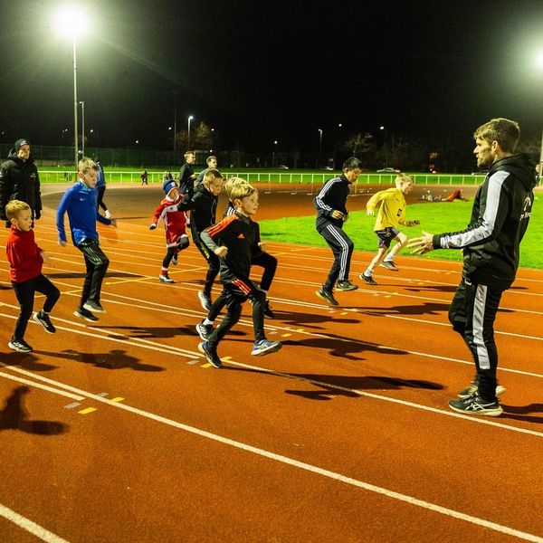 Children warming up on a track field at night with coach guidance.