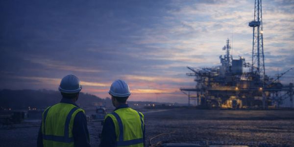 Two workers in safety gear observe an offshore oil platform at dusk.