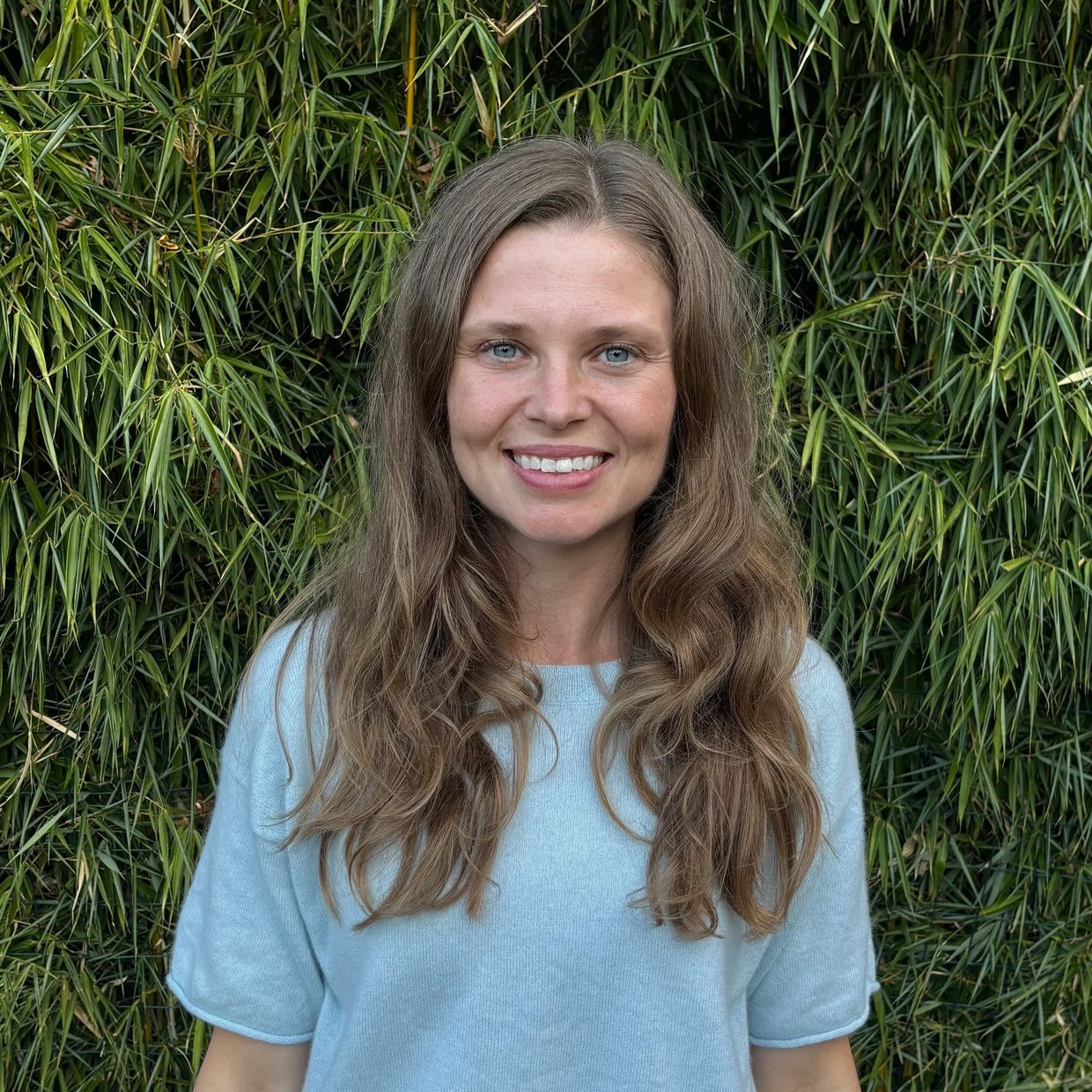 Smiling woman with long hair in front of green foliage.