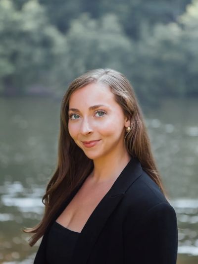 A confident woman in a black blazer poses outdoors by the water.
