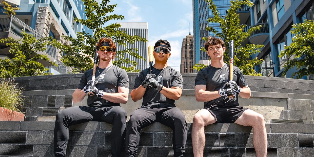 Three young men in sportswear with baseball bats sitting on a ledge in an urban area.