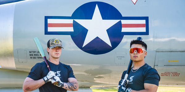 Two men pose with baseball bats in front of an aircraft with a U.S. Air Force insignia.