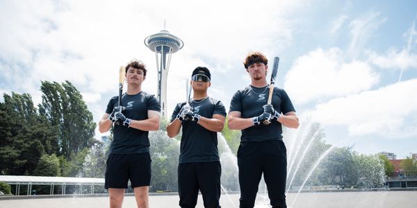 Three young athletes posing with baseball bats near a fountain and the Space Needle.