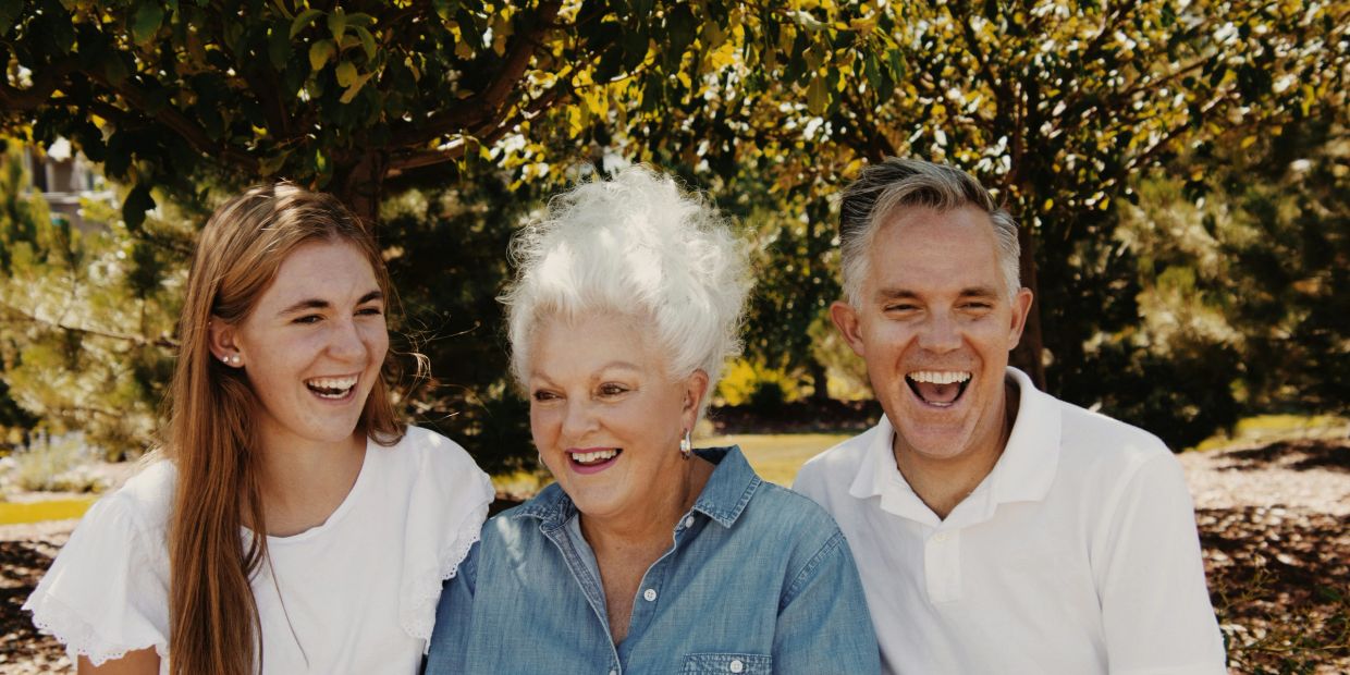 Three generations laughing outdoors under trees.