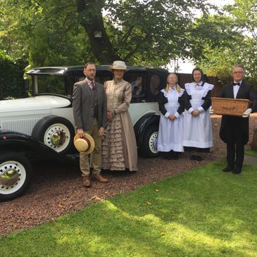 A couple in Victorian dress with 2 Maids and a butler in front of a vintage car in a garden.