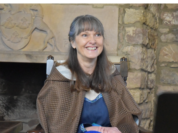 A seated woman in a castle, wearing a cloak smiling.