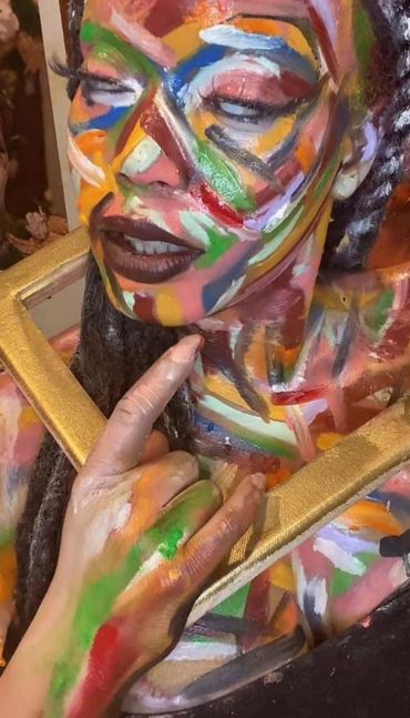 Person with colorful abstract face and body paint holding a golden frame.