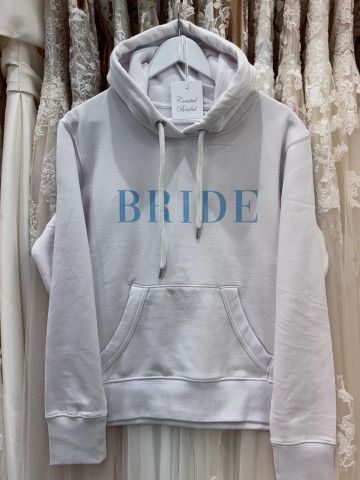 White hoodie with 'BRIDE' printed in blue, hanging among wedding dresses.