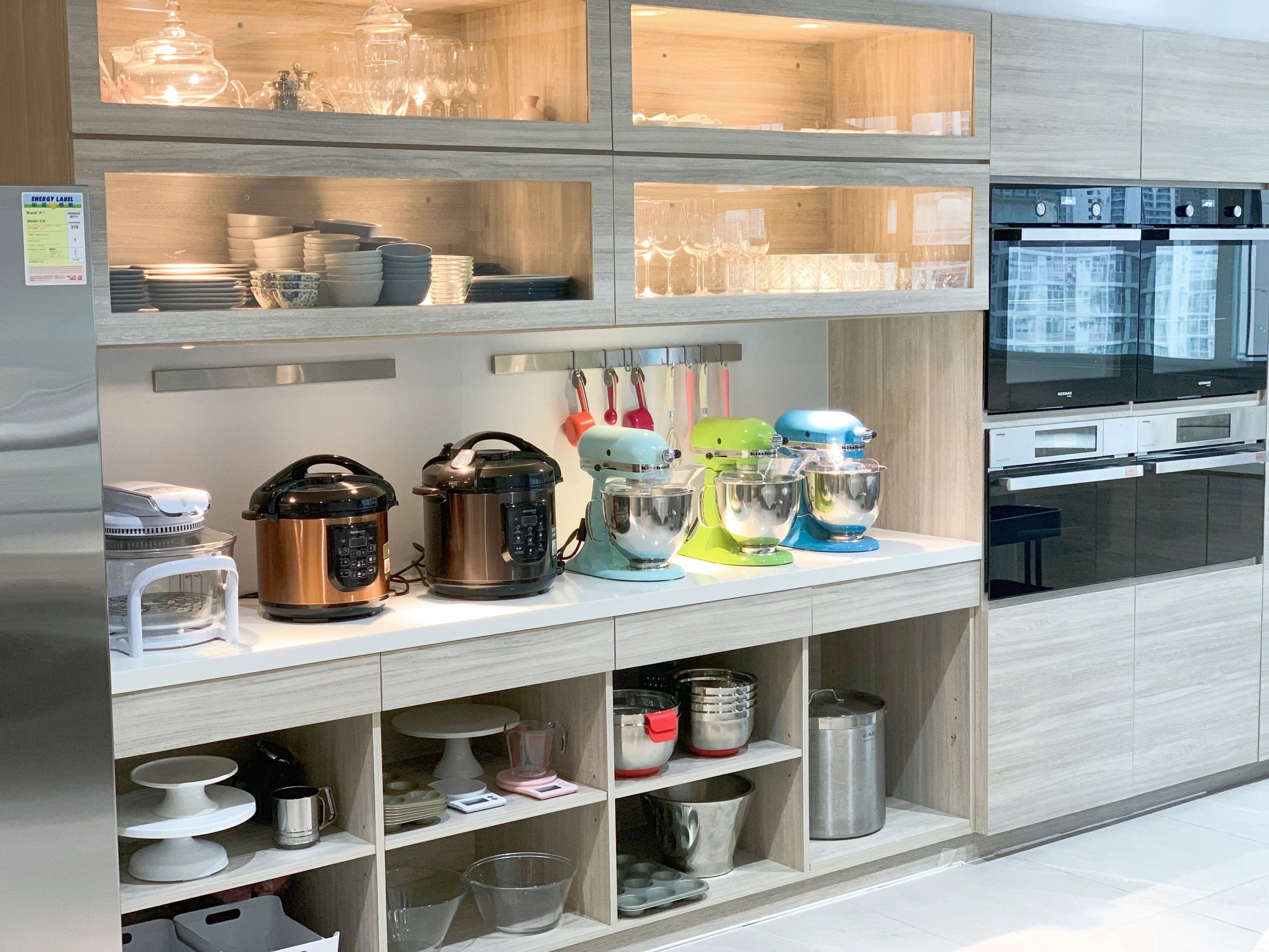 Modern kitchen with mixers, rice cookers, and glassware in wooden cabinets.