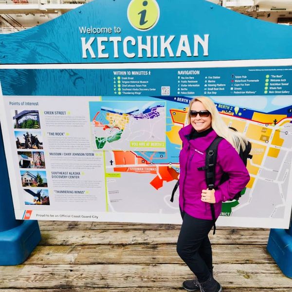 A woman in a purple jacket stands in front of a Ketchikan welcome sign and map.