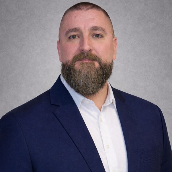 Man in a navy blazer with a full beard against a gray background.