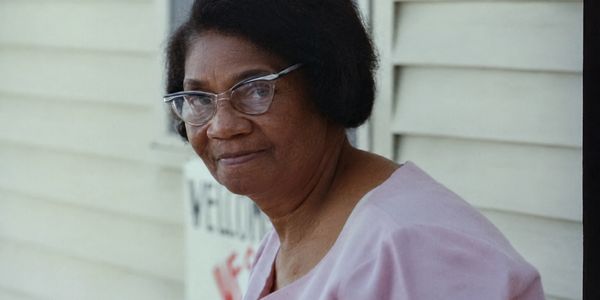 An elderly woman with glasses wearing a light pink blouse.