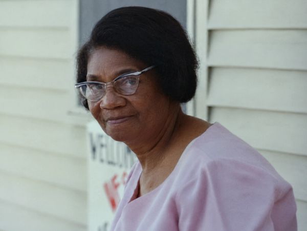 An elderly woman with glasses wearing a light pink blouse.