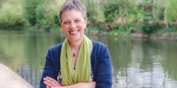 Jane Ide by a river. She has short greying dark hair, a blue top, bright green scarf and a big smile.