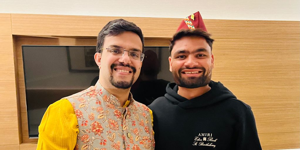 Two smiling men posing indoors, one in traditional attire, the other in casual wear with a festive hat.
