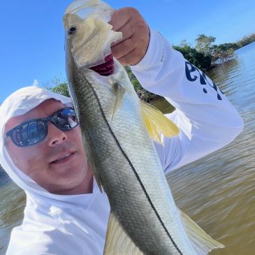 Man in white hoodie holding a large fish near a lake on a sunny day.