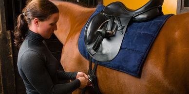 A woman adjusting a saddle on a brown horse inside a stable.