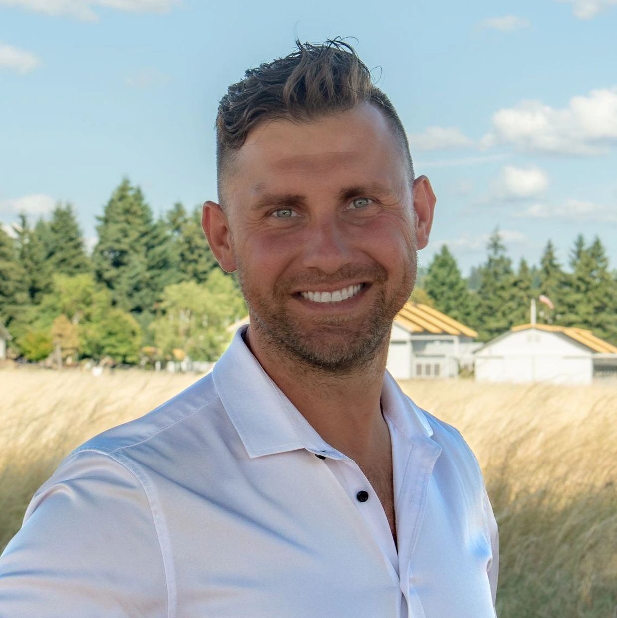 Smiling man in white shirt outdoors with trees and houses in background.