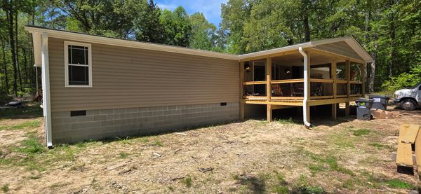 A beige mobile home with a covered wooden porch in a wooded area.