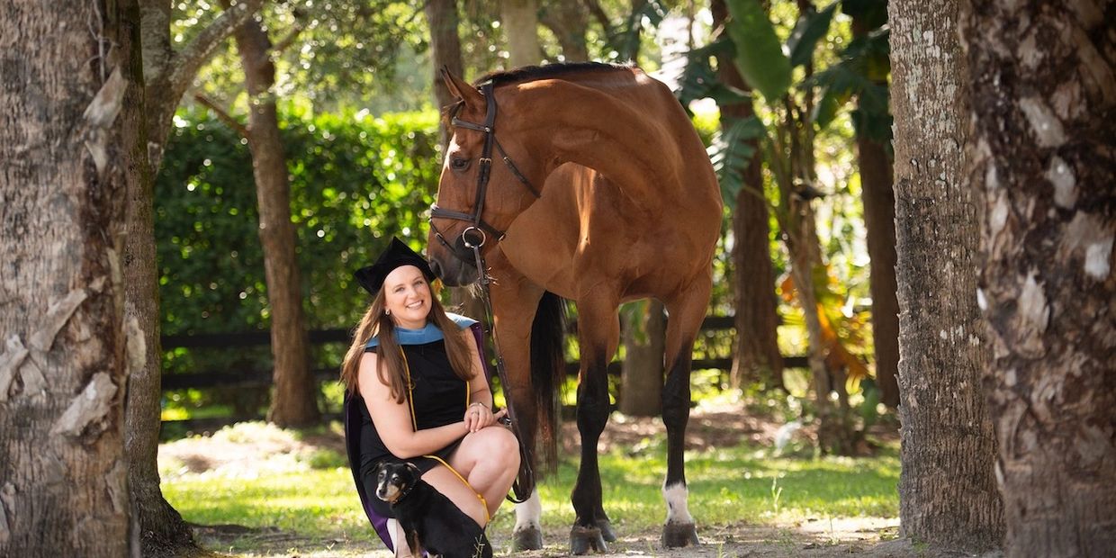 A graduate in cap and gown poses with her horse and dog in a sunlit forest.