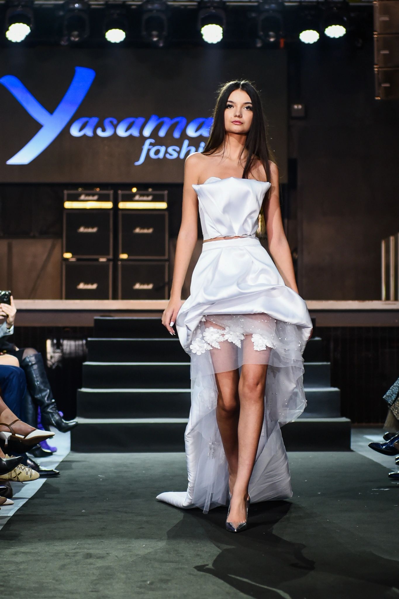 Model walking down the runway in a white, strapless dress with sheer floral detailing.