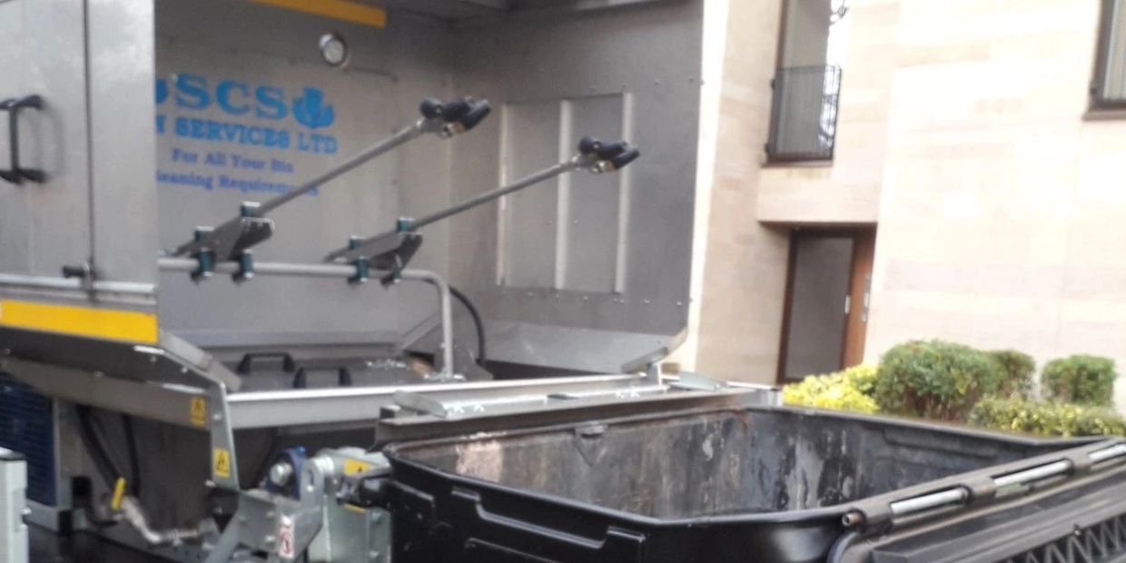 A garbage truck with an open black waste container beside it.