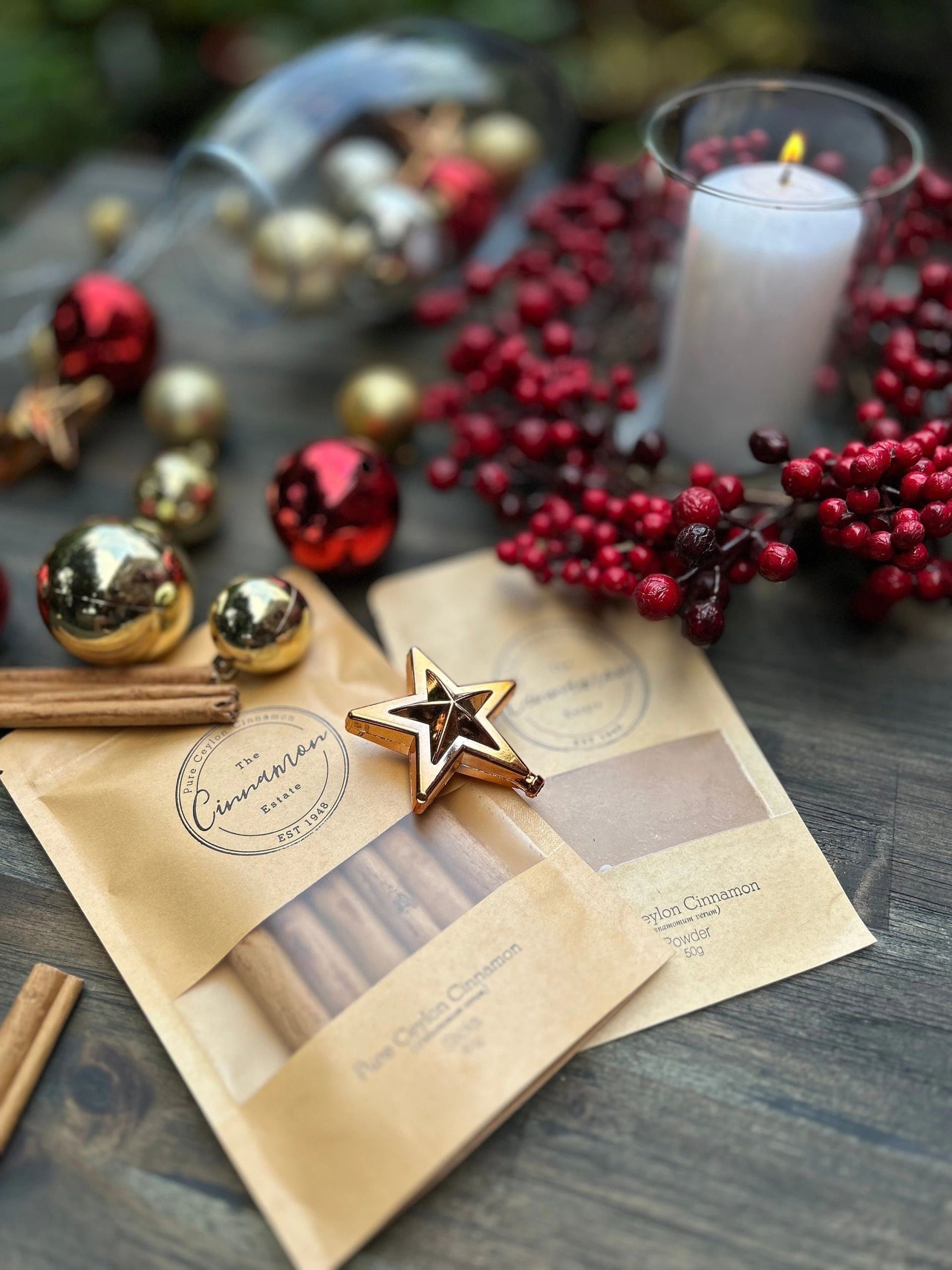 A kraft paper pouch of The Cinnamon Estate hand-rolled Ceylon cinnamon sticks, styled among red and gold Christmas ornaments, a berry wreath and a lit candle.