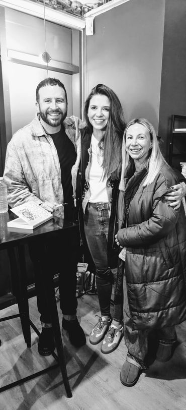 Three friends smiling and posing together indoors near a table with a book.
